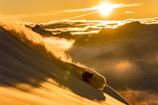 A person skiing in a cloud of snow above the clouds in French Alps at sunset.